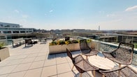 This rooftop terrace offers an exceptional outdoor experience set against the iconic Washington, D.C. skyline. The space is finished with large-format porcelain tiles that lend a clean, modern aesthetic underfoot, while glass railings along the perimeter preserve unobstructed panoramic views of the city — including a striking sightline to the Washington Monument in the distance. The terrace is thoughtfully furnished with contemporary laser-cut patterned chairs and round wooden tables, creating intimate seating areas ideal for gathering and conversation. Large metal planters filled with colorful flowering plants add warmth and a touch of nature to the urban setting, serving as both decorative accents and subtle dividers within the open-air layout. Additional seating arrangements are positioned throughout the terrace, accommodating a variety of social configurations. The open sky above and the expansive cityscape below combine to create a bright, airy ambiance that is both energizing and inviting. Whether used for casual relaxation or social events, this rooftop terrace delivers a premium urban outdoor experience with a commanding presence above the heart of the nation's capital.