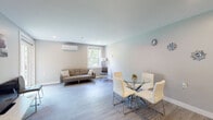 This single-floor unit showcases a clean, modern design with light gray walls, smooth ceilings, and consistent light-toned hardwood-style flooring throughout. The open-concept living and dining area is bright and airy, benefiting from multiple windows that frame lush green outdoor views, a sliding glass door leading directly to a private deck, and a wall-mounted mini-split HVAC unit for year-round climate control. The dining space accommodates a round glass-top table with contemporary chrome-legged chairs, while the living area offers comfortable seating alongside a second window view.

The galley-style kitchen is efficiently laid out with white shaker cabinetry, granite countertops, and a full suite of stainless steel appliances including a refrigerator, range, and over-the-range microwave, complemented by a stainless steel sink.

The bedroom is a calm, well-sized retreat with its own wall-mounted HVAC unit, a window overlooking greenery, recessed lighting, and ample space for a queen-sized bed and furnishings.

The bathroom is neatly finished with wood-look tile flooring, large-format gray tile surrounding a glass-enclosed walk-in shower, and clean white fixtures, creating a functional and contemporary space.