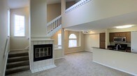 This two-story space makes an immediate impression with its dramatic double-height ceilings in the main living area, creating a sense of openness and grandeur. A striking tile-surround fireplace with glass doors serves as a focal point, anchoring the living room with both style and function. Natural light filters into the space through an arched window, brightening the neutral-toned carpeted floors and beige walls throughout.

A white-railed staircase ascends along one wall, leading to an open upper-level landing that overlooks the living area below, adding architectural depth and visual interest. The upper balcony features classic white balusters that complement the overall clean, traditional aesthetic of the space.

Adjacent to the living area, the kitchen is defined by a raised breakfast bar with a dark countertop, offering a natural transition between spaces. A wood-toned cabinet provides additional storage. Track lighting in this area ensures functional illumination. The overall ambiance is bright, airy, and well-proportioned, with the open floor plan and vertical space making the home feel expansive and inviting.
