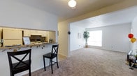This single-floor residence offers a thoughtfully designed open-concept layout that promotes a seamless flow between the kitchen and living areas. The kitchen features white cabinetry and is connected to the adjoining space via a convenient pass-through counter with bar seating, making it ideal for casual dining and entertaining. The counter is accented with a decorative garland, adding a welcoming touch to the space.

The living area is generously proportioned, finished with neutral wall-to-wall carpeting that lends warmth and comfort underfoot. A large window at the far end of the living space floods the room with abundant natural light, creating a bright and airy atmosphere. A potted indoor tree near the window adds a refreshing natural element to the interior.

Throughout the space, the walls are painted in a clean, neutral white tone, complemented by a textured ceiling finish that adds subtle visual interest. The overall aesthetic is light, functional, and inviting, with a practical layout that maximizes usable space. The smooth transition between the kitchen and living areas makes this residence well-suited for everyday living and comfortable hosting.