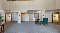 Located at 9950 Redwood St NW in Coon Rapids, Minnesota, this single-floor community space offers a thoughtfully designed layout that balances openness with comfort. The centerpiece is a dramatic living room featuring soaring vaulted ceilings and a striking floor-to-ceiling glass window wall that floods the space with natural light and provides views of the surrounding outdoors. An elegant chandelier hangs overhead, enhancing the grand atmosphere, while carpeted flooring and generous seating arrangements create a warm, inviting environment. Adjacent to the main living area, a library nook serves as a cozy retreat, lined with well-stocked bookshelves and anchored by a wood-framed gas fireplace beneath a wall-mounted television — an ideal setting for relaxed gathering and reading. A separate sitting area with a dining table and storage cabinet provides additional functionality. The hallway is spacious and well-lit, offering elevator access and a welcoming seating vignette with a cushioned chair and decorative floral arrangement. Throughout, neutral tones, plush carpeting, and consistent architectural detailing contribute to a cohesive, comfortable ambiance suited to community living.