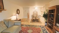 This single-floor space exudes a cozy and welcoming ambiance, thoughtfully designed to blend comfort with classic elegance. The open-plan layout seamlessly connects the living and dining areas, creating a sense of spaciousness and easy flow throughout.

The living area is anchored by a plush sofa and a richly patterned area rug in warm red and navy tones, adding a striking focal point to the room. A Tiffany-style lamp rests on a side table, casting a warm, decorative glow, while an ornate sunburst mirror adorns the wall, enhancing the room's traditional charm.

The dining area features a round glass-top table paired with wrought iron chairs, lending an elegant yet airy feel. A tall decorative baker's rack stands nearby, offering both visual interest and functional display space. A lush indoor plant adds a refreshing touch of greenery to the corner.

Light hardwood flooring runs throughout the space, complementing the warm, neutral wall tones and contributing to the bright, clean aesthetic. Upper glass-front cabinetry near the dining area hints at additional storage and display options. A ceiling fan overhead ensures year-round comfort. The entryway, with its step-up landing, provides a welcoming transition into this well-appointed and harmoniously styled space.
