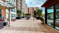 Located at 399 Main Ave in Norwalk, Connecticut, this outdoor amenity space is part of the Merritt River residential community — a modern, multi-building development defined by its striking contemporary architecture. The centerpiece of this level is a generously sized open-air courtyard framed by the community's tall residential towers, clad in a warm combination of brick red and neutral stucco facades with large teal-trimmed windows.

The courtyard is finished with large-format tiled pavers in a neutral palette, creating a clean and cohesive aesthetic throughout. Multiple seating zones are thoughtfully arranged across the space, including umbrella-shaded dining tables and chairs ideal for outdoor meals or socializing, as well as a lounge area featuring cushioned seating under a pergola with decorative columns. Large planters filled with mature trees, shrubs, and seasonal flowers are distributed throughout, adding natural greenery and visual interest.

Elegant lamp post signage bearing the "AJH" branding anchors the central walkway, lending a polished, curated feel to the environment. The lobby entrance, clearly marked with signage from Merritt River, features floor-to-ceiling glass walls that provide visibility into the building interior. The overall ambiance of the courtyard is open, bright, and welcoming, offering residents a comfortable and attractive outdoor retreat within the heart of the community.