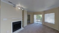 This single-floor residence offers a comfortable and functional living environment centered around a well-appointed main living area. The space is finished with plush wall-to-wall carpeting and warm neutral tones throughout, creating a cohesive and inviting atmosphere.

The living area serves as the focal point of the home, highlighted by a classic fireplace with a white mantle surround and an adjacent built-in shelving unit, ideal for display or storage. Track lighting installed along the ceiling provides adjustable, directed illumination across the room. Crown molding adds a refined architectural detail along the upper edges of the walls.

A structural column with decorative trim separates the living area from an adjoining room, providing a sense of defined space while maintaining an open flow. Large sliding glass doors dressed with vertical blinds open to an outdoor area, filling the interior with natural light and offering views of surrounding greenery. A secondary window further enhances the brightness of the space. The textured ceiling adds subtle depth to the overall interior finish, contributing to the room's understated, practical character.