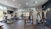 This single-floor fitness center is thoughtfully designed to support a comprehensive workout experience. The main exercise area is generously sized, accommodating a wide selection of cardio equipment including treadmills, elliptical trainers, and a step machine, all arranged to maximize usability and flow. A multi-station cable strength machine anchors the strength training zone, complemented by additional resistance equipment suited for a full-body workout. Large floor-to-ceiling mirrors line one wall, visually expanding the space and allowing users to monitor their form during exercise. The walls feature bold navy blue accents with athletic-themed graphic decals, adding an energetic, motivating ambiance to the neutral gray color scheme. Durable rubber flooring throughout provides both safety and comfort underfoot. Recessed lighting illuminates the space evenly, while oversized windows on multiple walls flood the interior with natural light and offer views of the surrounding greenery and an outdoor pool area. A wall-mounted flat-screen television provides entertainment during workouts. A separate transitional area near the entrance features speckled epoxy flooring, a navy accent wall, and a dedicated stretching or cool-down alcove, rounding out this well-appointed fitness facility.