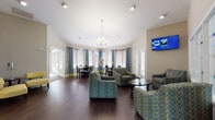 Located at 1000 Stonebrook Dr in Sanford, Florida, this single-floor community space is thoughtfully designed to provide a comfortable and inviting environment. The main lounge area features rich, dark hardwood flooring, vaulted ceilings, and an elegant chandelier as a focal point. Plush, patterned armchairs in teal and green tones are arranged around a central coffee table, creating an ideal setting for casual gatherings. A wall-mounted flat-screen display adds a modern touch to the space.

A separate dining area offers multiple glass-top tables surrounded by dark wood chairs with teal cushions, all bathed in natural light streaming through expansive windows dressed with two-tone teal and chartreuse drapes and wooden blinds. The bright, airy atmosphere of this area makes it well-suited for meetings or social events.

A cozy seating nook, accented with vibrant yellow chairs and contemporary artwork, provides a more intimate space for conversation. Adjacent to this area, a fully equipped kitchen features rich dark cabinetry, granite countertops, stainless steel appliances, and a peninsula with bar seating — ideal for catered events or resident use. The overall design palette of teal, yellow, and warm neutrals creates a cohesive, modern aesthetic throughout.