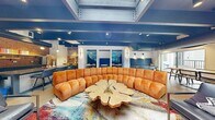 Located at 801 Camino del Rio in Durango, Colorado, this expansive single-floor lounge offers a generously sized, open-concept gathering space designed with a modern industrial aesthetic. Polished concrete floors, dark exposed ceiling beams, and visible ductwork establish a bold architectural character throughout, while a central skylight floods the space with natural light.

The main seating area anchors the lounge with a large curved sectional sofa in warm amber velvet, arranged around a striking live-edge slab coffee table atop a vibrant multicolor area rug. A built-in fireplace with a shiplap-clad surround and mounted TV adds a cozy focal point nearby. Accent chairs and additional seating arrangements provide flexible social spaces, complemented by intimate booth-style nooks upholstered in deep navy.

A full kitchen island with a corrugated metal base, dark countertops, and bar seating serves as the social hub of the space, backed by an exposed brick wall with open shelving and stainless steel appliances. Adjacent to the kitchen, a billiards table and high-top seating area cater to recreation, with large windows offering views into a connected fitness center. Mountain landscape murals add a distinctly Colorado character, while abundant windows and a glass-door entrance invite natural light and outdoor connection throughout.