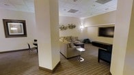This single-floor space is designed as a welcoming lounge or common area, offering a well-appointed environment suited for socializing or relaxing. The room features a neutral color palette of warm beige and cream tones on the walls, complemented by striped carpeting that adds visual texture underfoot. A drop ceiling with recessed lighting and wall-mounted sconces provides layered illumination throughout the space.

The seating area is anchored by a generously sized sectional sofa in a warm taupe fabric, accented with patterned throw pillows for comfort and style. A sleek, modern pedestal side table sits nearby, adding a contemporary touch. Opposite the seating area, a flat-screen television is mounted above a dark media console, creating a dedicated entertainment zone.

A structural column divides the space, adding architectural character while defining distinct functional zones. Adjacent to the column, a small bistro-style table and chairs offer an alternative seating option. Decorative circular metallic wall art adds an artistic focal point, enhancing the refined yet casual ambiance of the room. A potted plant near the seating area introduces a natural element, completing the inviting atmosphere.