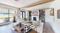 This well-appointed single-floor amenity space showcases a seamless blend of modern farmhouse and transitional design, creating a warm and inviting atmosphere throughout. Exposed wood ceiling beams add architectural character and visual warmth, while herringbone-patterned flooring flows through the main areas, anchoring the open layout with refined texture.

The central lounge area serves as the heart of the space, featuring a fireplace framed by built-in shelving and cabinetry, a wall-mounted display, and a mix of plaid and solid upholstered seating arranged around a round wood coffee table. Floor-to-ceiling black-framed windows and glass doors flood the area with natural light and offer views of lush outdoor greenery.

Adjacent to the lounge is a generously sized formal dining area finished in muted sage green, illuminated by elegant pendant lights with brass accents. A large wood dining table comfortably seats a group, complemented by upholstered chairs and a built-in window bench. Artwork and a sideboard complete the room's polished look.

A dedicated activity room provides flexible workspace with drafting tables, adjustable stools, built-in teal cabinetry, and a wall-mounted television. The wet bar area features sleek grey cabinetry, stone countertops, a wine refrigerator, and mirrored open shelving — ideal for entertaining. French doors provide direct access to outdoor areas.
