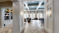 Located at 465 Elmcroft Blvd in Rockville, MD, this sophisticated single-floor community space offers a seamlessly connected living room, dining area, and kitchen across a generously sized open layout. The interior is immediately distinguished by striking coffered ceilings accented in teal, framed by crisp white molding that adds architectural depth and elegance throughout. Underfoot, a classic herringbone-patterned wood-look floor flows uniformly across the entire space, enhancing the sense of continuity and refined design. The living room is anchored by a white fireplace with a built-in bookshelf and media wall, complemented by teal upholstered seating and abundant natural light pouring in through floor-to-ceiling windows that offer views of an outdoor pool and patio area. Sheer drapery softens the light while maintaining the bright, airy ambiance. The kitchen features a large island with a crisp white countertop and navy blue base, surrounded by upholstered bar seating, white cabinetry, and integrated appliances. The dining area transitions naturally from the kitchen, creating a functional and stylish entertaining hub. Recessed lighting and thoughtfully placed wall art complete the polished, welcoming atmosphere of this beautifully appointed space.