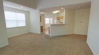 This two-floor residence is designed with an open, functional layout that promotes a sense of spaciousness across the main level. The living area features wall-to-wall carpeting in a neutral beige tone, creating a warm and comfortable foundation for the space. Recessed lighting complements the natural light streaming in through multiple windows, keeping the interior bright and welcoming.

The kitchen is positioned in an open-concept arrangement, separated from the living area by a raised breakfast bar peninsula with seating space — ideal for casual dining or entertaining. The kitchen is equipped with light oak cabinetry, a white refrigerator, a built-in microwave, and a tile or laminate floor that contrasts cleanly with the carpeted living zone. A flush ceiling light fixture provides focused illumination over the workspace.

Beyond the main living area, a connected secondary room — visible through an open doorway — features hardwood-style flooring and its own window, offering flexibility as a dining room or additional living space. The overall color palette is light and neutral throughout, with white walls and trim lending a clean, move-in-ready aesthetic across both levels.