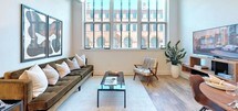 This stylish loft-style residence seamlessly blends industrial character with contemporary finishes across a single thoughtfully designed floor. Exposed ductwork runs along the ceilings, complementing the large, multi-pane factory-style windows that span entire walls, flooding every room with natural light and offering striking views of a neighboring historic brick building with arched detailing.

The open-plan living area features warm hardwood flooring and generous proportions, with the oversized windows creating a bright, airy atmosphere. The kitchen is outfitted with white cabinetry, granite countertops, stainless steel appliances including a gas range and French-door refrigerator, and a peninsula with bar seating beneath sleek pendant lighting.

Both bedrooms are well-sized, each benefiting from the dramatic window walls and hardwood floors. A dedicated flex room with a built-in desk and shelving provides an ideal home office or lounge space. The primary bathroom offers a glass-enclosed walk-in shower and a dark wood vanity with modern hardware, while the second bathroom features a soaking tub with shower combination and matching tile flooring. A walk-in closet with built-in rod storage and hardwood floors completes the primary suite, appointed with plush robes and slippers for a hotel-like touch.