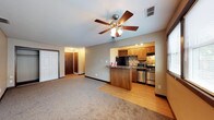 This single-floor space is designed with an open-concept layout that seamlessly connects the living area to the kitchen. The living area features plush carpeting underfoot and is topped by a ceiling fan with a built-in light fixture, providing both comfort and air circulation. Neutral-toned walls throughout create a clean, versatile backdrop.

The kitchen transitions from the carpeted living space via warm hardwood-style flooring and is defined by medium-toned wood cabinetry that extends to a dedicated wine rack above the refrigerator. A decorative tile backsplash adds visual texture behind the stainless steel appliances, which include a gas range, refrigerator, and microwave. The kitchen peninsula, finished with a dark granite-style countertop, serves as both a functional workspace and a natural divider between the kitchen and living areas.

A hallway visible in the background leads to additional rooms, flanked by what appears to be closet or utility access doors, suggesting practical storage solutions throughout the space. Recessed lighting in the kitchen, combined with the ceiling fan fixture in the living area, ensures the space is well-lit and functional.