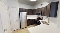 This single-floor space showcases a contemporary kitchen designed with both style and functionality in mind. The kitchen is outfitted with a full suite of stainless steel appliances, including a top-freezer refrigerator, an electric range with a smooth cooktop, and an over-the-range microwave, providing all the essentials for everyday cooking. Dark espresso-toned cabinetry lines the walls, offering generous upper and lower storage with sleek bar-pull hardware that contributes to a modern aesthetic. The countertops are finished in a light quartz with subtle veining, offering a clean contrast against the darker cabinetry. A stainless steel sink is integrated into the counter near a gooseneck faucet, adding a practical yet polished touch. Light wood-look flooring runs throughout, lending warmth to the space. The layout follows an efficient L-shaped configuration, maximizing workspace and movement. A peninsula counter visible in the foreground suggests additional seating or prep space. Neutral white walls and ceiling, combined with a flush-mount light fixture and a ceiling vent, complete the crisp, well-maintained interior.