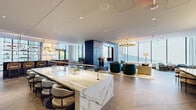 Located at 1909 Walnut Street in Philadelphia's vibrant Rittenhouse Square neighborhood, this single-floor amenity lounge is a masterclass in contemporary luxury design. The open-plan layout flows seamlessly across herringbone-patterned hardwood floors, connecting a series of distinct yet cohesive gathering spaces bathed in abundant natural light from expansive, floor-to-ceiling windows that frame striking views of the surrounding cityscape.

At the heart of the space sits a commanding marble-topped island bar, its dramatic white and gold veining set against a backdrop of teal-painted walls adorned with abstract gold brushstroke accents. Sleek white bar seating lines the island, while a fully stocked back bar with pendant lighting creates an inviting focal point. An adjacent lounge area features plush seating in rich jewel tones — deep teal, cognac, and warm grey — anchored beneath a statement brass chandelier and complemented by a bold blue tile fireplace surround.

Intimate seating alcoves positioned along the window line invite relaxed conversation, while a dedicated dining area with built-in bench seating and a long communal table offers a more structured gathering option. A powder room showcases artisan-level detail with bespoke teal and gold wallcovering and a marble vanity. French doors lead to an outdoor terrace, further enhancing the connection between interior elegance and the dynamic Philadelphia skyline.