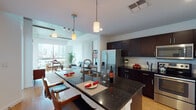 This modern urban residence blends industrial design elements with contemporary finishes to create a stylish and functional living space. The open-concept layout connects the kitchen, dining, and living areas seamlessly, all anchored by warm hardwood flooring throughout.

The kitchen is a standout feature, outfitted with rich dark cabinetry, white subway tile backsplash, stainless steel appliances — including a French door refrigerator, electric range, and built-in microwave — and a spacious island with a dark granite countertop that doubles as a breakfast bar. An in-unit washer is conveniently tucked just off the kitchen.

The living and dining area is bathed in natural light from expansive floor-to-ceiling windows that frame impressive city skyline views. Exposed concrete ceiling details lend an industrial character, while a textured concrete accent wall adds depth and visual interest to the space.

The bedroom continues the industrial-modern aesthetic with exposed concrete ceilings, large windows, and direct access to a private balcony overlooking the cityscape, filling the room with abundant natural light.

The bathroom features a full tub-shower combination with clean white tile surround, dark wood vanity with granite countertop, and crisp white tile flooring, completing this well-appointed urban residence.