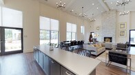 This beautifully appointed clubhouse offers a refined, open-concept layout that seamlessly blends functionality with upscale design. The space is anchored by dramatic, soaring ceilings adorned with exposed beamed detailing and a beadboard finish, creating an airy and expansive atmosphere throughout. Multiple contemporary chandeliers with sputnik-style arms provide stylish ambient lighting that complements the natural light pouring in through the floor-to-ceiling roller shade windows, offering views of the surrounding community.

The kitchen area features a generous island with a crisp white quartz countertop and sleek dark cabinetry, ideal for catering and social gatherings. Adjacent to the kitchen, a large dining area accommodates a substantial table with ample seating for group events and meetings. The lounge area transitions naturally from the dining space, offering comfortable seating arrangements set atop a plush area rug, centered around a striking stone fireplace that serves as a warm focal point. A sculptural decorative piece adds an artistic touch to the overall ambiance. The hardwood-style flooring unifies the entire space, tying together the neutral, contemporary color palette of warm whites, grays, and natural wood tones for a welcoming and polished communal environment.
