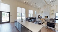 This beautifully appointed clubhouse offers a refined, open-concept layout that seamlessly blends functionality with upscale design. The space is anchored by dramatic, soaring ceilings adorned with exposed beamed detailing and a beadboard finish, creating an airy and expansive atmosphere throughout. Multiple contemporary chandeliers with sputnik-style arms provide stylish ambient lighting that complements the natural light pouring in through the floor-to-ceiling roller shade windows, offering views of the surrounding community.

The kitchen area features a generous island with a crisp white quartz countertop and sleek dark cabinetry, ideal for catering and social gatherings. Adjacent to the kitchen, a large dining area accommodates a substantial table with ample seating for group events and meetings. The lounge area transitions naturally from the dining space, offering comfortable seating arrangements set atop a plush area rug, centered around a striking stone fireplace that serves as a warm focal point. A sculptural decorative piece adds an artistic touch to the overall ambiance. The hardwood-style flooring unifies the entire space, tying together the neutral, contemporary color palette of warm whites, grays, and natural wood tones for a welcoming and polished communal environment.