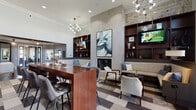 Located at 2800 Pontiac Dr in Aurora, IL, this single-floor community clubhouse is thoughtfully designed to offer residents a refined and comfortable gathering space. The main lounge commands attention with soaring ceilings, contemporary sputnik-style chandeliers, and a dramatic floor-to-ceiling tile fireplace flanked by dark wood built-in shelving with integrated flat-screen televisions. A large-format abstract artwork anchors the focal wall, adding an artistic touch to the sophisticated interior. Seating is arranged to encourage both social interaction and relaxed lounging, featuring a mix of upholstered benches, accent chairs, and a substantial wooden bar-height table with leather seating. A cozy nook adjacent to the fireplace offers built-in banquette seating beside a window with outdoor views. The kitchen area is outfitted with white quartz countertops, rich wood cabinetry, frosted glass upper cabinets, pendant lighting, and a coffee station, providing a fully functional communal space. A dedicated business center offers workstation setups with computer monitors in a quiet, separately defined area, accented by large-format tile flooring and a statement mirror. The entry foyer welcomes visitors with leather armchairs, curated artwork, and tall glass doors that flood the space with natural light.