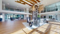 Located at 5201 Congress Ave in Boca Raton, Florida, this single-story commercial space delivers an impressive blend of modern design and refined elegance. The grand lobby serves as the centerpiece, boasting soaring double-height ceilings adorned with decorative exposed wood beam accents and striking woven pendant light fixtures that add a warm, organic contrast to the crisp white architecture. Wide-plank hardwood flooring extends throughout, anchoring the expansive open layout.

Floor-to-ceiling glass walls and large curtain-wall windows flood the interior with natural light while offering views of the surrounding Florida landscape. A glass-railed mezzanine level overlooks the main floor, connected by a sleek marble-clad staircase. Marble and stone column details punctuate the space, complementing the contemporary aesthetic.

Distinct zones include a welcoming reception area featuring a wood-and-marble desk, carpeted lounge areas with curated furnishings, and glass-enclosed meeting or club rooms with interior views. Bold contemporary artwork decorates the walls throughout. The overall ambiance is polished yet approachable, combining upscale architectural details with an open, airy atmosphere suited for a dynamic professional or amenity-driven environment.