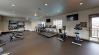 This fitness center offers a comprehensive workout environment designed to support a wide range of exercise routines. The space is laid out across a single open floor, finished with durable dark rubber flooring throughout and illuminated by recessed ceiling lighting supplemented by natural light from multiple windows that provide views of the surrounding exterior.

The cardio zone is well-stocked with several treadmills, elliptical trainers, and stationary bikes, arranged to make efficient use of the open floor plan. Multiple wall-mounted flat-screen televisions are positioned throughout the space for entertainment during workouts.

The strength training area features a multi-station cable and weight machine setup, along with a dedicated free weights section equipped with a tiered dumbbell rack holding a full range of weights. Adjustable flat benches are positioned nearby for versatile use. A separate alcove area houses additional selectorized strength machines, offering a quieter zone for targeted muscle training.

A standout feature is the TRX functional training wall unit, which includes suspension trainers, stability balls, medicine balls, and resistance bands — providing a complete functional fitness station. The neutral-toned walls and clean, organized layout contribute to an efficient and motivating atmosphere suited for a full-body workout experience.