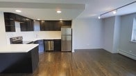 This single-floor space offers a thoughtfully designed open-concept layout that seamlessly connects the kitchen and living areas, creating a spacious and functional environment. The kitchen is a standout feature, boasting dark espresso-toned cabinetry that provides ample storage, complemented by a classic white subway tile backsplash that adds a clean, contemporary contrast. Stainless steel appliances — including a full-size refrigerator and dishwasher — contribute a polished, modern aesthetic. Light countertops balance the rich tones of the cabinetry, while recessed lighting overhead ensures the kitchen is well-illuminated. Rich, dark hardwood flooring runs throughout the space, adding warmth and continuity from the kitchen into the open living area. The living space is generously sized, offering flexibility for a variety of furniture arrangements, and is equipped with track lighting that adds both function and style. Neutral gray walls provide a versatile backdrop that suits a range of décor styles. The overall ambiance is modern and inviting, with a cohesive design language that balances dark and light tones to create a comfortable, stylish living environment.