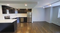 This single-floor space offers a thoughtfully designed open-concept layout that seamlessly connects the kitchen and living areas, creating a spacious and functional environment. The kitchen is a standout feature, boasting dark espresso-toned cabinetry that provides ample storage, complemented by a classic white subway tile backsplash that adds a clean, contemporary contrast. Stainless steel appliances — including a full-size refrigerator and dishwasher — contribute a polished, modern aesthetic. Light countertops balance the rich tones of the cabinetry, while recessed lighting overhead ensures the kitchen is well-illuminated. Rich, dark hardwood flooring runs throughout the space, adding warmth and continuity from the kitchen into the open living area. The living space is generously sized, offering flexibility for a variety of furniture arrangements, and is equipped with track lighting that adds both function and style. Neutral gray walls provide a versatile backdrop that suits a range of décor styles. The overall ambiance is modern and inviting, with a cohesive design language that balances dark and light tones to create a comfortable, stylish living environment.