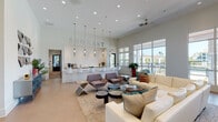 The 500 East clubhouse lounge is a thoughtfully designed, single-floor communal space that blends contemporary aesthetics with functional comfort. The open-plan layout is anchored by soaring ceilings that amplify the sense of space, while recessed lighting and ceiling fans are evenly distributed throughout to maintain a bright, comfortable atmosphere.

The lounge area is generously proportioned, featuring a large cream sectional sofa complemented by accent chairs in warm taupe tones, arranged around a cluster of dark, varied-height coffee tables atop a neutral area rug. Colorful artwork and decorative accents introduce vibrant pops of color against the predominantly white and light-toned interior. Large-format tile flooring in a subtle hexagonal pattern adds a refined textural element underfoot.

Adjacent to the lounge, a prominent bar-style counter finished in crisp white stretches along one wall, accented by a textured white tile backsplash and suspended cylindrical pendant lights. White bar stools line the counter, which is equipped with a coffee station and bar accessories, providing a casual gathering point for residents.

Floor-to-ceiling gridded windows along one side of the space flood the interior with natural light and offer unobstructed views of the outdoor surroundings, creating a seamless connection between the interior and exterior environment.