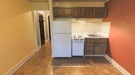 This single-floor space offers a practical and functional layout with a cohesive, no-frills design aesthetic. The kitchen serves as a notable focal point, featuring dark-stained wood cabinetry both above and below the countertops, providing ample storage. White appliances, including a full-size refrigerator and a coil-burner range with oven, complement the light-colored countertop and single-basin stainless steel sink. A light-toned tile backsplash lines the walls behind the cooking and prep areas, adding a subtle visual detail. The kitchen floor is dressed in a distinctive brick-patterned tile, which creates a clear and charming delineation from the warm hardwood-style flooring that extends throughout the rest of the space. A hallway leading from the kitchen area provides access to additional rooms, with doorways visible in the background suggesting a bedroom and bathroom. The warm yellow-toned walls throughout contribute to a cozy, inviting ambiance. The space is straightforward in its design, prioritizing functionality while maintaining a welcoming character through its warm color palette and traditional materials.
