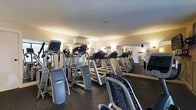 This fitness center offers a dedicated space for cardiovascular and general exercise, thoughtfully designed to accommodate a range of workout routines. The facility is outfitted with a substantial lineup of cardio machines, including treadmills and elliptical trainers, arranged in orderly rows to maximize usable floor space. The flooring features a dark, rubberized surface that provides both durability and traction underfoot — a practical choice suited to high-traffic exercise environments. Full-length mirrors line the walls, visually expanding the space while allowing users to monitor their form during workouts. Warm, ambient wall sconces complement the overhead lighting to create a well-lit atmosphere throughout. A wall-mounted air conditioning unit ensures comfortable temperature regulation regardless of the intensity of activity. The smooth, light-toned ceiling adds a clean, finished look to the interior. A glimpse through the mirrored reflection suggests an adjacent area or entryway, indicating that the fitness center may be part of a larger facility. Overall, the gym presents a clean, organized, and functional environment well-suited for consistent, effective exercise.