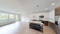 This contemporary single-floor unit showcases a clean, modern design with a seamless open-concept layout connecting the kitchen and living areas. The kitchen is well-appointed with a two-tone cabinetry scheme — crisp white upper cabinets paired with warm wood-toned lower cabinets — complemented by a full suite of stainless steel appliances, including a refrigerator, range, microwave, and dishwasher. A freestanding kitchen island with a stainless steel countertop and built-in shelving provides additional prep space and storage flexibility. Light wood-look flooring runs consistently throughout the unit, lending warmth and cohesion to the interior. The open living area is bright and airy, bathed in natural light through expansive sliding glass doors and large windows that frame views of lush green landscaping. The bedroom continues the unit's clean aesthetic with neutral walls, wood-look flooring, a generous window with outdoor views, and direct access to a closet. The bathroom features a warm wood vanity with a white countertop, a large mirror, and neutral tile flooring, maintaining the unit's contemporary and understated style. Recessed lighting throughout ensures a well-lit environment, while the overall design emphasizes simplicity, functionality, and modern living.
