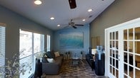 This single-floor space offers a thoughtfully designed interior that blends comfort with a coastal aesthetic. The living area serves as the centerpiece of the home, showcasing a soothing blue-toned color palette that sets a relaxed and inviting atmosphere throughout. A striking large-scale ocean wave artwork anchors one wall, reinforcing the space's coastal theme and drawing the eye immediately upon entry.

The living room is well-appointed with a plush gray sectional sofa accented with decorative throw pillows, positioned around a glass-topped coffee table that rests on a patterned area rug. A sleek media console lines the opposite wall, providing both functional storage and entertainment space. Natural light filters into the room through multiple windows dressed with horizontal blinds, contributing to the bright and airy feel of the space.

Overhead, recessed lighting is thoughtfully distributed across the ceiling, complemented by a ceiling fan with wood-toned blades that adds both style and practical airflow. The ceiling features a subtle texture, and the overall layout is open and efficiently arranged. Decorative plants and accent pieces are placed throughout, adding organic touches that enhance the room's welcoming character. The space is cohesive, polished, and designed with livability in mind.