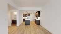 This single-floor residence showcases a clean, contemporary design with light hardwood-style flooring flowing throughout the main living areas. The open-concept kitchen serves as the centerpiece of the space, anchored by a substantial island with a white quartz countertop, an integrated sink, and stylish pendant lighting overhead. Rich dark-toned cabinetry lines the walls, providing generous storage, while stainless steel appliances—including a refrigerator, range, and microwave—complete the well-appointed cooking space. The kitchen transitions seamlessly into a spacious living area, which is bathed in natural light from large sliding glass doors that lead to a private balcony. The living space offers an open, airy feel with white walls and recessed lighting throughout. The bedroom features plush neutral carpeting and a window that allows natural light to filter in. The bathroom is outfitted with a textured dark vanity, white quartz countertop, a wide mirror with strip lighting above, and a separate shower enclosure. Overall, the space presents a cohesive modern aesthetic with a neutral palette and quality finishes.