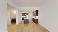 This single-floor residence showcases a clean, contemporary design with light hardwood-style flooring flowing throughout the main living areas. The open-concept kitchen serves as the centerpiece of the space, anchored by a substantial island with a white quartz countertop, an integrated sink, and stylish pendant lighting overhead. Rich dark-toned cabinetry lines the walls, providing generous storage, while stainless steel appliances—including a refrigerator, range, and microwave—complete the well-appointed cooking space. The kitchen transitions seamlessly into a spacious living area, which is bathed in natural light from large sliding glass doors that lead to a private balcony. The living space offers an open, airy feel with white walls and recessed lighting throughout. The bedroom features plush neutral carpeting and a window that allows natural light to filter in. The bathroom is outfitted with a textured dark vanity, white quartz countertop, a wide mirror with strip lighting above, and a separate shower enclosure. Overall, the space presents a cohesive modern aesthetic with a neutral palette and quality finishes.