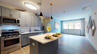 Located at 100 Oakview Dr in Trumbull, Connecticut, this single-floor residence offers a thoughtfully designed layout with modern finishes and a cohesive aesthetic throughout. The open-concept kitchen is a standout feature, showcasing gray shaker-style cabinetry, a white subway tile backsplash, stainless steel appliances including a gas range, microwave, and French door refrigerator, and a generous center island with a crisp white countertop illuminated by stylish pendant lighting. Wood-look flooring flows seamlessly from the kitchen into the adjacent living area, which is bathed in natural light from large windows overlooking lush greenery. The living space offers a calm, airy atmosphere with soft neutral wall tones and room for generous furnishings. The primary bathroom features a dual-sink vanity with gray cabinetry, a large mirror, light tile flooring, and a walk-in glass-enclosed shower with floor-to-ceiling tile surround. A second bathroom provides a tiled soaking tub and shower combination with neutral finishes. The floor plan, visible in the dollhouse view, reveals a well-proportioned layout with clearly defined rooms connected by convenient corridors, making efficient use of the available space throughout this well-appointed single-level home.