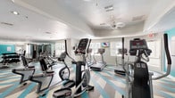 This fitness center is a thoughtfully designed, single-floor facility that offers a comprehensive workout environment. The space is immediately distinguished by its striking geometric-patterned flooring, featuring bold teal, gray, and white chevron shapes that lend a modern, energetic aesthetic throughout the room.

The cardio zone is well-appointed with multiple treadmills, elliptical trainers, and a recumbent bike, several of which face a wall-mounted flat-screen television for entertainment during workouts. A ceiling fan and recessed lighting ensure comfort and visibility throughout the space. Natural light filters in through multiple windows, enhancing the open, airy atmosphere.

The strength training area is equally comprehensive, housing a multi-station cable machine, a dumbbell rack with a full range of free weights, a weight bench, and a barbell station. A large wall of floor-to-ceiling mirrors runs along one side, visually expanding the space and allowing users to monitor their form.

A vibrant teal accent wall anchors one end of the room, providing visual contrast and housing accessories such as a wall-mounted exercise mat. The facility also includes a dedicated open area with medicine balls, suitable for functional training and stretching. Direct exterior access through a glazed door connects the fitness center to the surrounding outdoor environment.