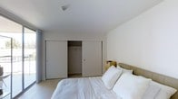 This single-floor space features a bedroom designed with a clean, minimalist aesthetic. The room is finished in soft, neutral whites and light grays, creating a calm and restful atmosphere. Smooth white walls and a flat white ceiling contribute to the airy, uncluttered feel of the space.

A standout storage feature is the wide, double sliding-door closet that spans a significant portion of one wall. With one panel open, the interior reveals a hanging rod and ample storage depth, offering practical organization for clothing and personal items. The sliding door design maximizes usable floor space by eliminating the need for swing clearance.

The bedroom accommodates a full-sized bed dressed in crisp white bedding, complemented by a neutral upholstered headboard and a woven storage basket on the floor beside it, adding a subtle natural texture to the otherwise understated palette. Light-toned flooring runs throughout, reinforcing the room's bright and cohesive design.

Overall, the space presents a functional and well-organized bedroom with a modern, no-frills design approach suited to those who appreciate simplicity and practicality in their living environment.