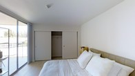 This single-floor space features a bedroom designed with a clean, minimalist aesthetic. The room is finished in soft, neutral whites and light grays, creating a calm and restful atmosphere. Smooth white walls and a flat white ceiling contribute to the airy, uncluttered feel of the space.

A standout storage feature is the wide, double sliding-door closet that spans a significant portion of one wall. With one panel open, the interior reveals a hanging rod and ample storage depth, offering practical organization for clothing and personal items. The sliding door design maximizes usable floor space by eliminating the need for swing clearance.

The bedroom accommodates a full-sized bed dressed in crisp white bedding, complemented by a neutral upholstered headboard and a woven storage basket on the floor beside it, adding a subtle natural texture to the otherwise understated palette. Light-toned flooring runs throughout, reinforcing the room's bright and cohesive design.

Overall, the space presents a functional and well-organized bedroom with a modern, no-frills design approach suited to those who appreciate simplicity and practicality in their living environment.