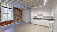This studio in Memphis, TN offers a compelling blend of industrial character and functional design. The open-concept main living area immediately draws the eye with its striking exposed brick wall, which spans the full length of the space and adds rich texture and historic charm. Overhead, painted open-beam ceilings with visible structural joists reinforce the industrial aesthetic, while linear fluorescent lighting illuminates the polished concrete floors below.

The kitchen is thoughtfully laid out in an L-shaped configuration, featuring ample white cabinetry reaching toward the ceiling for generous storage, gray laminate countertops, and a full suite of white appliances including a refrigerator, range with overhead microwave, dishwasher, and double sink. The clean, bright cabinetry provides a crisp contrast to the rawer elements found throughout the space.

A window along the brick wall invites natural light and offers views of the surrounding urban streetscape, connecting the interior to the downtown Memphis environment.

The bathroom is well-appointed with a white vanity, gray countertop, and a glass-enclosed tiled shower featuring colorful mosaic tile accents in purple and teal tones, adding a distinct visual focal point. Together, these elements make this studio a distinctive urban retreat with standout architectural character.