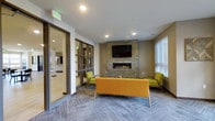 This multi-story residential community is designed with a contemporary aesthetic, combining blue and neutral-toned siding, brick accents, and large windows throughout its exterior. The building wraps around a spacious central courtyard finished with decorative paver pathways, raised planter beds, and colorful outdoor seating, creating an inviting communal gathering space. Mature landscaping and sculptural planters add greenery and visual interest throughout the courtyard.

A dedicated pool terrace offers a resort-style experience, with a swimming pool surrounded by a wide concrete deck furnished with shaded dining tables and lounge seating beneath large umbrellas. An additional outdoor patio area, covered by a slatted wood pergola, provides a sunlit yet sheltered space with tile flooring and ample seating for relaxing or dining al fresco.

Inside, the communal lounge features a floor-to-ceiling stacked stone fireplace with a mounted flat-screen television above it, complemented by bold yellow and green accent furnishings set atop patterned carpet. Glass-paneled sliding doors connect this lounge to adjacent common areas, enhancing the open, connected feel of the interior amenity spaces.