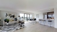 This single-floor space is designed around an open-concept layout that integrates the living, dining, and kitchen areas into one cohesive environment. The most striking feature is the expansive floor-to-ceiling sliding glass doors that span the width of the main living area, flooding the interior with abundant natural light and providing direct access to a private balcony with open city views. The light-toned hardwood flooring runs throughout, reinforcing the airy, contemporary aesthetic.

The living area is anchored by a large area rug with a geometric hexagonal pattern in grey and white tones, complementing the neutral color palette of white walls and modern furnishings. Abstract wall art adds visual interest. The dining area sits adjacent, featuring a sleek table and modern chairs, positioned to take full advantage of the natural light and balcony views.

The kitchen is partially open to the main space, separated by a breakfast bar with bar seating. White cabinetry and stainless steel appliances contribute to the clean, modern look. Recessed lighting and contemporary wall décor complete the polished, cohesive design of this well-appointed space.