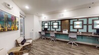 This single-floor space is designed as a contemporary business center and communal lounge, combining functionality with polished interior design. The layout is organized into distinct zones that serve different purposes while maintaining a cohesive aesthetic throughout.

The workstation area is a standout feature, offering a long granite-topped counter lined with multiple ergonomic task chairs and computer workstations. The backdrop wall is a striking design element, composed of a geometric pattern in teal and dark wood tones, accented by square wall-mounted sconces that cast a warm, layered glow. A shared printer is conveniently integrated into the workspace.

Adjacent to the workstation zone, a small private meeting room accommodates a four-seat dining-height table and upholstered chairs. Bold, large-format artwork — including a vibrant Route 66-themed piece — decorates the walls, while linen drape panels soften the room.

The lounge area serves as a welcoming transitional space, furnished with gold-framed cream accent chairs, a wood cube side table, and colorful cityscape artwork displayed prominently on white walls. Glass-panel doors connect this area to neighboring rooms, enhancing openness and visibility. Throughout, a multi-toned striped carpet grounds the space with a contemporary yet warm finish.