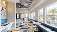 Located at Integra TC in Daytona Beach, Florida, this thoughtfully designed tech hub and lounge space offers a refined, contemporary environment ideal for work and relaxation. The single-floor layout encompasses three distinct functional zones that flow seamlessly into one another.

The main lounge area is anchored by a striking slatted ceiling feature with integrated linear lighting, adding architectural depth and visual interest overhead. Large double-hung windows line the walls, flooding the space with abundant natural light and offering pleasant views of palm trees and the surrounding community. A built-in navy banquette runs along the window wall, paired with sleek marble-topped bistro tables and modern white accent chairs, creating an inviting social or work setting.

A relaxed seating nook features a wall-mounted flat-screen TV, framed abstract artwork in blue tones, and a pair of plush lounge chairs arranged around a mirrored accent table — ideal for casual collaboration or downtime. The bold geometric area rug in navy, gold, blue, and gray unifies the space with a vibrant, modern energy.

A semi-private workspace, separated by black-framed glass partitions, offers a quiet retreat with a marble-topped desk, textured wall coverings, and a view-facing window. A built-in blue cabinetry unit with a printer station serves practical office needs. Throughout, the neutral warm palette, polished finishes, and curated furnishings create a cohesive, upscale ambiance.