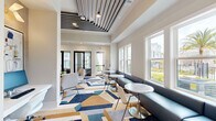 Located at Integra TC in Daytona Beach, Florida, this thoughtfully designed tech hub and lounge space offers a refined, contemporary environment ideal for work and relaxation. The single-floor layout encompasses three distinct functional zones that flow seamlessly into one another.

The main lounge area is anchored by a striking slatted ceiling feature with integrated linear lighting, adding architectural depth and visual interest overhead. Large double-hung windows line the walls, flooding the space with abundant natural light and offering pleasant views of palm trees and the surrounding community. A built-in navy banquette runs along the window wall, paired with sleek marble-topped bistro tables and modern white accent chairs, creating an inviting social or work setting.

A relaxed seating nook features a wall-mounted flat-screen TV, framed abstract artwork in blue tones, and a pair of plush lounge chairs arranged around a mirrored accent table — ideal for casual collaboration or downtime. The bold geometric area rug in navy, gold, blue, and gray unifies the space with a vibrant, modern energy.

A semi-private workspace, separated by black-framed glass partitions, offers a quiet retreat with a marble-topped desk, textured wall coverings, and a view-facing window. A built-in blue cabinetry unit with a printer station serves practical office needs. Throughout, the neutral warm palette, polished finishes, and curated furnishings create a cohesive, upscale ambiance.