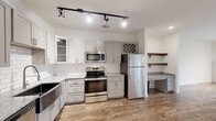 Located at 1000 Sweet Almond Dr in Holly Springs, NC, this well-appointed one-bedroom, one-bathroom residence offers a thoughtfully designed single-floor layout. The kitchen is a standout feature, showcasing gray shaker-style cabinetry, granite countertops, a white subway tile backsplash, a stainless steel farmhouse sink, and a full suite of stainless steel appliances including a range, microwave, dishwasher, and refrigerator. A built-in wine rack and open wood shelving add both character and functionality. Light wood-look flooring flows through the main living areas, complemented by track lighting that adds a modern touch. The living room opens directly to a private balcony through a glass door, inviting natural light and outdoor views. The bedroom is carpeted for comfort, features a ceiling fan, large windows with garden views, and direct access to the bathroom. The bathroom includes a tub-shower combination and wood-look flooring. A dedicated closet with wire shelving systems provides organized storage. Clean white walls and neutral finishes throughout create a bright, cohesive aesthetic suited for everyday living.