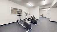 Located at 125 Broad St in Weymouth, Massachusetts, this fitness center offers a thoughtfully organized, single-floor layout designed to accommodate a wide range of workout routines. The space features clean white walls, durable rubber-composite flooring, and bright fluorescent lighting throughout, creating a functional and welcoming exercise environment.

The cardio area is equipped with elliptical trainers and a stationary cycle, providing ample options for aerobic workouts. Adjacent to this zone, a dedicated strength training section houses treadmills and a cable/functional trainer machine, along with a flat utility bench, supporting both resistance and full-body training exercises.

A free weights area features a well-stocked dumbbell rack and an adjustable weight bench, ideal for targeted strength work. Nearby, a functional training nook contains kettlebells in multiple sizes, medicine balls, resistance tools, and stability balls, catering to dynamic and circuit-style training.

An open, multipurpose area with rubber flooring and a water fountain provides space for stretching, floor exercises, or cool-down routines. Interior windows connect the various zones, promoting visibility across the facility. The space also includes a private door, likely leading to restroom or utility access. Overall, this fitness center is well-equipped and efficiently laid out to serve a broad range of fitness needs.