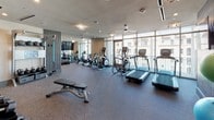 Located at 2902 Carlisle St in Dallas's vibrant Uptown district, this fitness center is a comprehensively equipped facility designed to support a wide range of workout routines. The single-level space is thoughtfully divided into distinct workout zones, each outfitted with professional-grade equipment.

The cardio area is a standout feature, lined with floor-to-ceiling windows that flood the room with natural light and offer open views of the surrounding cityscape. Treadmills, elliptical machines, and stationary cycling bikes are arranged along this windowed wall, creating an energizing environment. Ceiling fans maintain airflow throughout, and a wall-mounted flat-screen TV provides entertainment during workouts.

The strength and free-weight area houses a full rack of dumbbells in a range of sizes, an adjustable bench, and a dedicated functional training station equipped with kettlebells, medicine balls, resistance bands, and suspension trainers. A large mirror expands the visual depth of the space, while framed artwork and textured accent walls contribute a polished aesthetic. Yoga mats and accessories are neatly organized for group or individual stretching and flexibility work. Recessed lighting and neutral tones throughout create a clean, professional atmosphere well-suited for focused training.