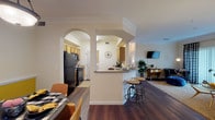Located in San Antonio's Hardy Oak area, this single-story residence offers a thoughtfully designed open-concept layout that seamlessly connects the main living areas. Dark hardwood flooring flows throughout, complemented by crisp white crown molding and decorative arched doorways that lend a refined architectural character to the interior.

The living room is bright and welcoming, featuring large sliding glass doors dressed with bold patterned curtains that allow generous natural light to fill the space. The kitchen is efficiently laid out with warm wood cabinetry, black appliances including a refrigerator, range, and built-in microwave, and a peninsula bar with seating that opens directly to the dining area — ideal for casual entertaining.

Two bedrooms provide comfortable retreats, each with plush carpeting and ample closet space. The primary bedroom is particularly serene, with soft neutral tones and good proportions. The primary bathroom is well-equipped with rich wood cabinetry, an extended vanity with a large mirror, hardwood-style flooring, and a spacious walk-in closet with built-in wire shelving. A dedicated walk-in closet off the bathroom adds further storage convenience.

Overall, the residence presents a warm and cohesive aesthetic, blending classic architectural details with functional, everyday living spaces.