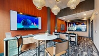 Located at 100 S 48th St in Phoenix, Arizona, this thoughtfully designed single-floor lounge space combines modern aesthetics with a warm, inviting ambiance across two distinct areas.

The living room is a standout gathering space anchored by a bold terracotta-toned accent wall featuring a distinctive scalloped texture pattern. A large-scale abstract painting adds artistic depth, while a plush olive-green sectional sofa with layered throw pillows provides comfortable seating. A multi-panel wall-mounted display system serves as a striking focal point, making this area ideal for entertainment and communal viewing. Patterned carpeting and a round dark wood coffee table complete the cozy, curated atmosphere.

The dining area offers a bright, open contrast with expansive floor-to-ceiling sliding glass windows that flood the room with natural light and frame views of the surrounding desert landscaping, including palm trees and gravel gardens. Warm wood-paneled walls add richness to the space, while a round bistro-style table paired with sleek metal-framed chairs creates an intimate setting. A wall-mounted television and a compact counter with storage provide additional functionality. The patterned area rug ties both zones together, reinforcing the space's cohesive and contemporary design identity.
