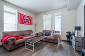 This single-floor unit offers a thoughtfully designed layout encompassing multiple bedrooms, a full kitchen, living area, and bathrooms across a generously sized floor plan. The kitchen is a standout feature, showcasing rich dark-wood cabinetry, granite countertops, stainless steel appliances including a gas range, built-in microwave, and refrigerator, all illuminated by stylish track lighting and pendant fixtures. A breakfast bar with seating extends from the kitchen, creating a seamless transition into the living and dining area. The living space is bright and open, benefiting from large floor-to-ceiling windows dressed with vertical blinds that allow for ample natural light. Durable wood-style flooring runs throughout the common areas and bedrooms, adding warmth and continuity. The bedrooms are comfortably sized, each featuring windows with blinds and open-concept closet or shelving areas. The hallway connects the rooms efficiently, lined with neutral-toned walls and dark door trim for a clean, modern contrast. Bathrooms include a walk-in shower stall with a curtain enclosure and chrome fixtures, as well as a separate lavatory. The overall design aesthetic is modern and functional, with neutral walls and consistent finishes throughout.